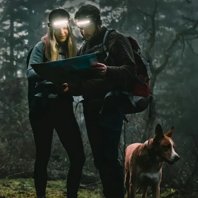 Zwei Wanderer mit eingeschalteten Stirnlampen prüfen im dunklen Wald eine Papierkarte, ihr Hund steht daneben